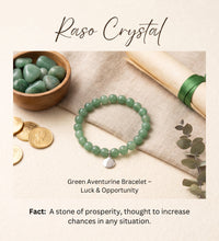 A green aventurine beaded bracelet with a silver heart charm is displayed on a beige fabric surface, surrounded by a wooden bowl of polished green stones, gold coins, a rolled parchment with a green ribbon, and eucalyptus leaves. The text 'Raso Crystal' is at the top, and 'Green Aventurine Bracelet - Luck & Opportunity' with a fact about the stone is at the bottom.