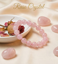 A pink crystal beaded bracelet arranged on a soft cream fabric backdrop, with a small white dish holding dried rose petals and a dried rose, plus several loose polished pink crystal stones. The words 'Raso Crystal' appear at the top.