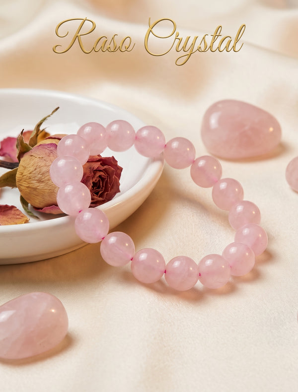 A pink crystal beaded bracelet arranged on a soft cream fabric backdrop, with a small white dish holding dried rose petals and a dried rose, plus several loose polished pink crystal stones. The words 'Raso Crystal' appear at the top.